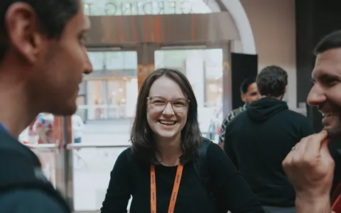 A smiling conference attendee with glasses and an orange lanyard chatting with others.